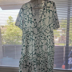 Vit blommig klänning med grönt mönster - Superfin vit klänning med grönt blommigt mönster och v-ringning. Modellen har korta ärmar och en lös, flowy passform som ger en chill vibe. Perfekt för varma dagar när du vill känna dig både snygg och bekväm.