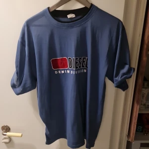 Blå t-shirt från Diesel - Säljer en blå t-shirt från Diesel med tryck 'Denim Division' på framsidan. T-shirten har en klassisk rund hals och korta ärmar. Perfekt för en avslappnad stil.