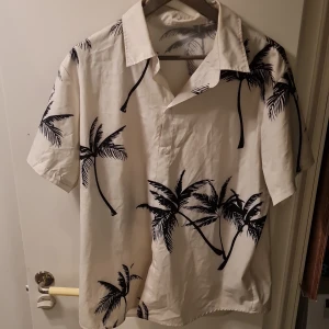 Vit kortärmad skjorta med palmmönster - Snygg vit skjorta med korta ärmar och ett stilrent svart palmmönster. Perfekt för en avslappnad sommarlook. Skjortan har en klassisk krage och knappar framtill.