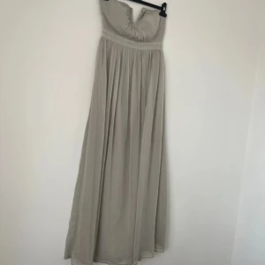 Grå axelbandslös balklänning  - Elegant grå axelbandslös långklänning med vacker drapering och pärldetaljer vid midjan. Perfekt för en stilfull kväll eller speciellt tillfälle.