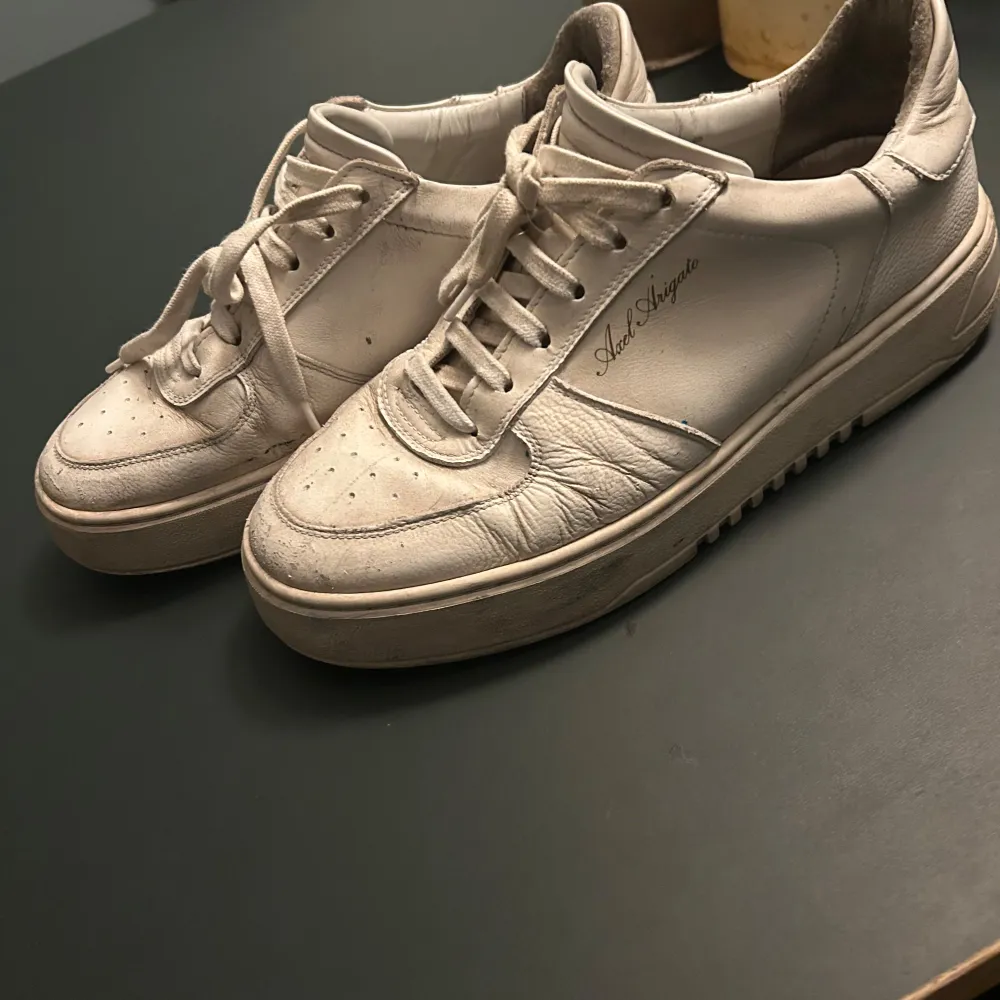 Snygga vita sneakers från Axel Arigato med klassisk snörning och rund tå. Skorna har en stilren design med märkets logga på sidan och en robust sula med texten 'ARIGATO' under. Perfekta för en casual look.. Kengät.