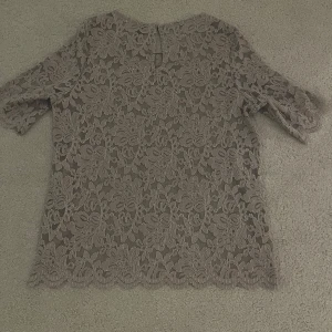 Grå Beige spetsblus😍 - Säljer en elegant beige spetsblus med korta ärmar och rund hals. Blusen har ett vackert blommönster och en knapp i nacken för enkel påklädning. Perfekt för en stilren look. Supersöt nu till våren 😍