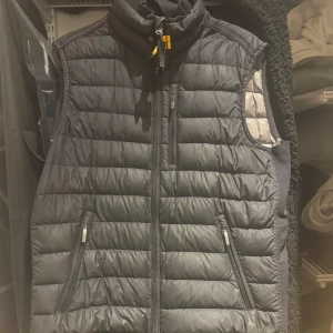 Svart dunväst från Parajumpers - Säljer en snygg svart dunväst från Parajumpers. Västen har en dragkedja framtill och praktiska fickor med dragkedja. Perfekt för lager-på-lager under kyliga dagar. Den har en stilren design med märkets logga på baksidan. Barn storlek med passar XS-S dam oxå