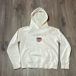 Vit hoodie från GANT - Säljer en stilren vit hoodie från GANT med en broderad logga och ett emblem på bröstet. Tröjan har en klassisk känguruficka och ribbade muddar vid ärmslut och nederkant. Perfekt för en avslappnad stil. 🧢