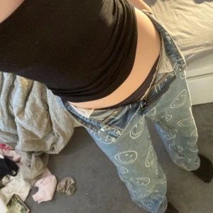 Blå baggy jeans med smiley-mönster  - Säljer ett par coola blå jeans från H&M med ett unikt smiley-mönster. Byxorna har en normal passform och är perfekta för att ge din outfit en rolig touch. De är tillverkade i jeansmaterial och har en klassisk dragkedja och knapp framtill. Ger en baggy effekt. Högmidjade fast om man är smal längst ner blir det lågmidjade