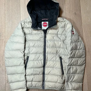 Beige pufferjacka från Colmar - Säljer en snygg beige pufferjacka från Colmar med huva och dragkedja. Jackan har ett quiltat mönster och två praktiska fickor med dragkedja. Perfekt för kyliga dagar!