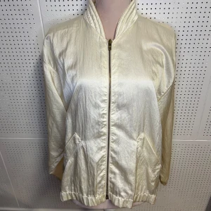 Beige bomberjacka med dragkedja - Snygg beige bomberjacka med dragkedja framtill och praktiska fickor. Jackan har en stilren design med långärmade ärmar och en lätt glansig finish. Perfekt för en trendig look. En liten fläck finns på högra armen, se bild. 