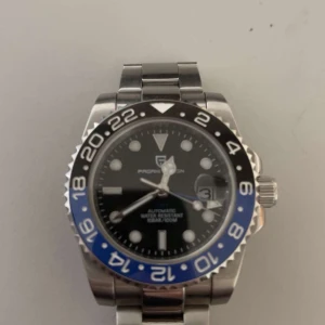 Pagani klocka - Säljer en automatisk klocka från Pagani Design med en stilren svart urtavla och blå-svart bezel. Klockan har en robust silverfärgad länkarmband och tydliga markeringar för timmar och datum. Perfekt för den som söker en kombination av stil och funktion. Går ner i pris vid snabb affär