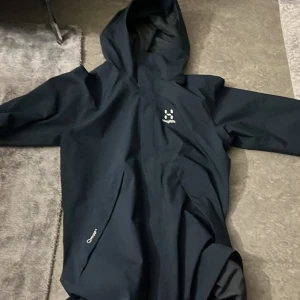 Marinblå regnjacka från Haglöfs - Säljer en svart regnjacka från Haglöfs med huva och dragkedja. Jackan har en stilren design och är perfekt för regniga dagar. Den är tillverkad i ett syntetmaterial som håller dig torr och bekväm. Storleken är S, den är inprincip ny, använd max 2 gånger!