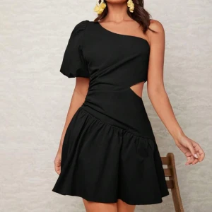 Svart oneshoulder klänning - Snygg svart oneshoulder klänning med en unik cut-out detalj i midjan. Klänningen har en volangdetalj vid kjolen och är perfekt för en kväll ute. Den korta längden ger en trendig look.