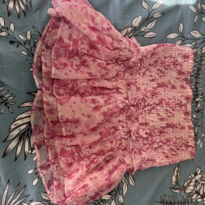 Volangkjol - Söt rosa kortkjol med blommigt mönster och volangdetaljer. Perfekt för en somrig look. Kjolen har en elastisk midja för bekväm passform.