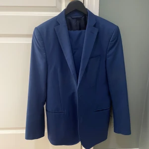 Blå kostym från J.Lindeberg - Snygg blå kostym från J.Lindeberg med elegant passform. Kostym har två knappar framtill och är fodrad med ett glansigt tyg. Perfekt för en stilren look. Uppskattar storleken till en 48 eller M.
