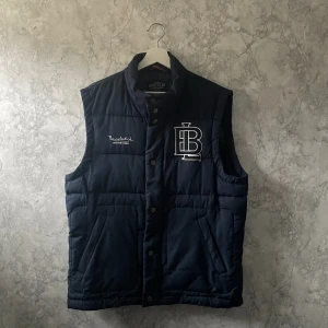 Mörkblå väst från Bondelid - Säljer en snygg mörkblå väst från Bondelid med broderad logga på bröstet. Västen har knappar och en praktisk dragkedja framtill. Perfekt för lager-på-lager under kyliga dagar.