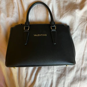 Svart handväska från Valentino  - Elegant svart handväska från Valentino med guldfärgade detaljer och dubbla dragkedjor. Väskan har två handtag och en stabil botten med metallfötter. Perfekt för att bära dina dagliga nödvändigheter med stil.använd några gånger ingen skada