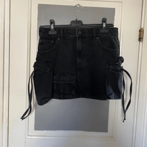 Svart jeanskjol med fickor - Snygg svart jeanskjol med stora fickor på sidorna och knytband. Perfekt för en cool och avslappnad stil. Kjolen har en knapp och dragkedja framtill.