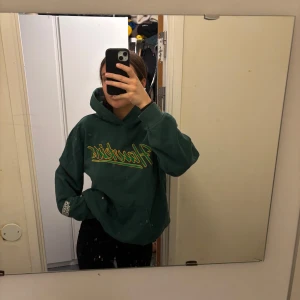 Grön hoodie med text - Säljer en grön hoodie med texten 'Hawkins' i gult och vitt på framsidan. Hoodien har en klassisk passform med en stor ficka framtill och långa ärmar. Perfekt för en avslappnad stil.