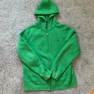 Grön hoodie från Polo Ralph Lauren - Säljer en snygg och ny grön hoodie från Polo Ralph Lauren med dragkedja och klassisk logga på bröstet. Perfekt för en avslappnad stil. Den har långa ärmar och en praktisk huva. Nypris: 1150 kr