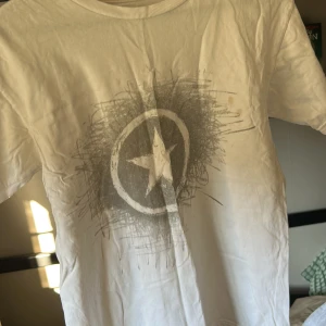 Vit t-shirt med stjärntryck från Converse - Säljer en vit t-shirt från Converse med ett coolt stjärntryck i grått på framsidan. T-shirten är kortärmad och tillverkad i 100% bomull, vilket gör den skön och bekväm att bära. Perfekt för en avslappnad stil!