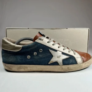 Snygga sneakers med stjärndetalj - Unika sneakers från Golden Goose i en cool kombination av blått jeansmaterial och brunt skinn. Skorna har en iögonfallande stjärndetalj i silver på sidan och klassisk snörning. Perfekta för en avslappnad stil med en touch av lyx. Box medföljer, ny pris 4999kr