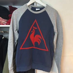 Blå och grå sweatshirt med röd tupp - Snygg sweatshirt från Le Coq Sportif i blått och grått med en stor röd tupp-logga på framsidan. Tröjan har långa ärmar och rund halsringning. Perfekt för en avslappnad stil.