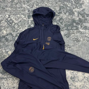 Nike mörkblå dress PSG - Säljer en mörkblå hoodie och matchande byxor från Nike med guldfärgade detaljer och logotyp. Hoodien har en praktisk dragkedja och en bekväm passform. Perfekt för sport och avslappnade dagar.