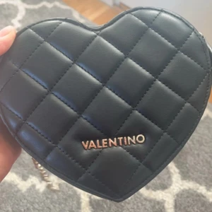 Axelväska från Valentino - Snygg svart hjärtformad axelväska från Valentino med quiltad design och kedjerem. Väskan har en dragkedja upptill och är perfekt för att bära dina nödvändigheter med stil. Den eleganta designen gör den till ett utmärkt val för speciella tillfällen.Den är i perfekt skick utvändigt och ser ut som ny! Endast lite smuts inuti, men inget som påverkar användningen. Öppen för förhandling.