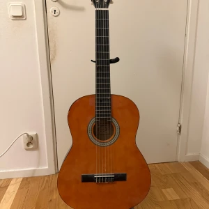 Klassisk akustisk gitarr - Hej! Säljer en klassisk akustisk gitarr med en vacker träfinish. Gitarren har sex strängar och en dekorativ rosett runt ljudhålet. Perfekt för både nybörjare och erfarna spelare som vill ha ett pålitligt instrument. Strängarna är relativt nya och gitarren har knappt använts. Fodral och ställ finns kommer med på köpet! 