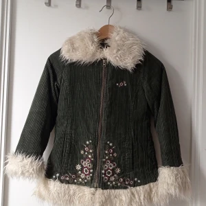 Grön manchesterjacka från Lindex - Säljer en grön manchesterjacka från Lindex med fluffig vit fuskpäls vid krage, ärmslut och nederkant. Jackan har en dragkedja framtill och fina broderade blommor i rött och vitt. Perfekt för kyliga dagar! Storlek 134. tyvärr är dragkedja lite trög men fungerar fortfarande att öppna och stänga. 