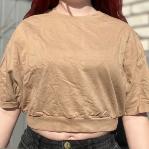 Beige croppad t-shirt - Säljer en beige croppad t-shirt med korta ärmar. Perfekt för en avslappnad stil. T-shirten har en rund halsringning och är gjord i ett mjukt material.