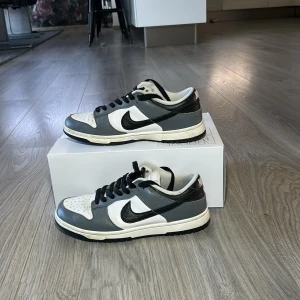 Nike Dunk i svart, vit och grå - Snygga Nike Dunk sneakers i svart, vit och grå färgkombination. Skorna har en klassisk design med snörning och rund tå. Perfekta för en stilren look.