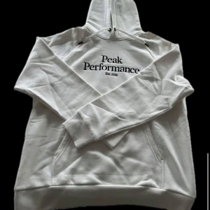 Vit hoodie från Peak Performance - Säljer en stilren vit hoodie från Peak Performance med svart broderad logga på bröstet. Tröjan har en klassisk design med känguruficka och justerbar huva. Perfekt för en avslappnad stil.