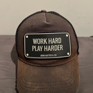 Svart keps från John Hatter & Co - Snygg svart keps med svart nätpanel och ett coolt märke framtill med texten 'Work Hard Play Harder'. Perfekt för en avslappnad stil.
