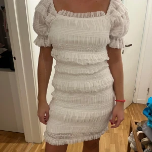 Vit klänning  - Säljer en söt vit klänning från Dry Lake med volanger och puffiga kortärmar. Klänningen har en smickrande passform och är perfekt för sommarens festligheter. Den har en elastisk struktur som ger en bekväm passform. Prislappen är kvar och den är aldrig använd! 