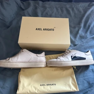 Vita sneakers från Axel Arigato - Snygga vita sneakers från Axel Arigato med en stilren design och en svart fågeldetalj på sidan. Skorna har snörning och en rund tå, vilket ger en klassisk look. Perfekta för en avslappnad stil.
