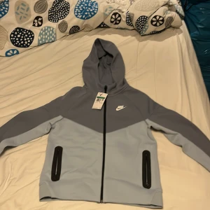 Grå hoodie från Nike - Säljer en stilren grå/ljusblå hoodie från Nike med dragkedja och huva. Jackan har två svarta fickor med dragkedjor framtill och en vit Nike-logga på bröstet. Perfekt för en avslappnad stil. Storlek XL (kids), helt ny aldrig använt