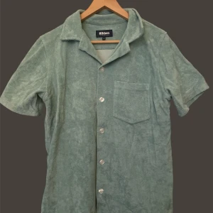 Nikben  - Snygg grön kortärmad skjorta i frotté från Nikben. Skjortan har en klassisk krage och knappar framtill samt en bröstficka. Perfekt för en avslappnad stil.