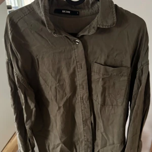 Olivgrön skjorta från Bik Bok - Säljer en olivgrön skjorta från Bik Bok med klassisk krage och bröstficka. Skjortan har långa ärmar och knappar framtill. Perfekt för en stilren look.