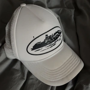 Corteiz keps - Snygg vit keps med svart tryck av ett fartyg på framsidan. Kepsen har ett nätmaterial på baksidan för bättre ventilation och en justerbar rem för perfekt passform.