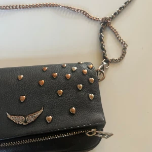 Svart axelväska med hjärtan och kedja - nano rock bag från Zadig & Voltaire med dekorativa hjärtan och en kedja som axelrem. Väskan har en dragkedja och ett unikt vingmotiv i silver. hjärtarna har missfärgats men inget man tänker på, utöver det är den i mycket bra skick❤️ kan gå ner i pris vid snabb handel!!