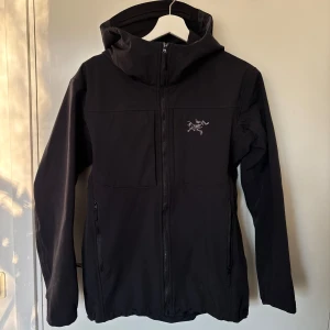 Svart jacka från Arc'teryx - Snygg svart jacka från Arc'teryx med dragkedja och broderad logga på bröstet. Jackan har en stilren design med långa ärmar och praktiska fickor med dragkedja. Har också två fickor vid bröstet och anpass bar luva. Perfekt för utomhusaktiviteter.