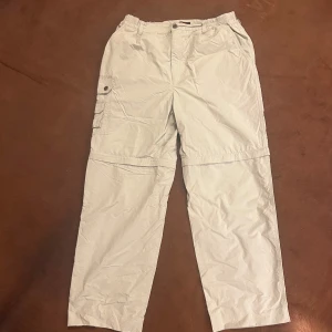 Beige cargopants med avtagbara ben - Säljer ett par praktiska beige cargopants från Tillander med avtagbara ben. Byxorna har flera fickor med knappar och en svensk flagga som detalj. Perfekta för utomhusaktiviteter och äventyr.