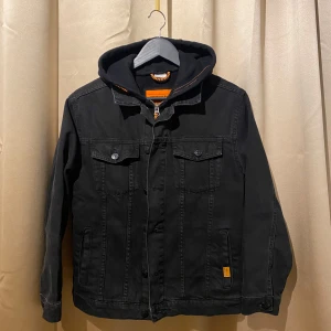 Svart jeansjacka med huva - Snygg svart jeansjacka med avtagbar huva och orange detaljer. Jackan har knappar framtill och två bröstfickor med knappstängning. Perfekt för en casual look.