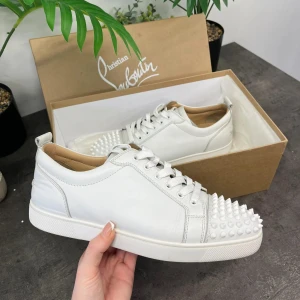 Vita sneakers med nitar från Christian Louboutin - Snygga vita sneakers från Christian Louboutin med coola nitar på tån och den ikoniska röda sulan. Skorna har snörning och är tillverkade i skinn, vilket ger en lyxig känsla. Perfekta för att ge din outfit en edgy touch.