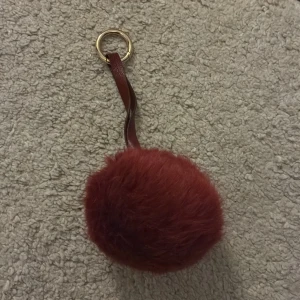 Vinröd pälsboll nyckelring - Säljer en vinröd pälsboll nyckelring med en guldfärgad ring och röd läderrem. Perfekt som accessoar till din väska eller nyckelknippa.