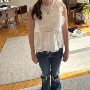 Vit blus med volangdetaljer - Säljer en söt vit blus med volangdetaljer och knapp i nacken. Blusen är ärmlös och har en luftig passform, perfekt för varma dagar.