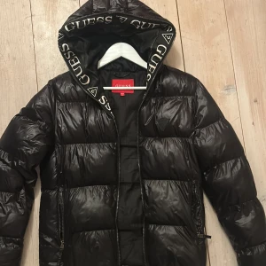 Svart pufferjacka från Guess - Säljer en snygg svart pufferjacka från Guess med logotyp på huvan. Skit snygg och skön. 
