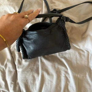 Väska massimo dutti - Snygg svart mini axelväska i skinn med flätade detaljer på sidorna. Väskan har en justerbar axelrem och en praktisk dragkedja upptill. Perfekt för att bära dina nödvändigheter med stil. 