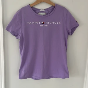 Lila t-shirt från Tommy Hilfiger - Säljer en snygg lila t-shirt från Tommy Hilfiger i bomull. T-shirten har korta ärmar och ett tryck med märkesloggan och 'Est. 1985' på framsidan. Frakt ingår inte i priset!