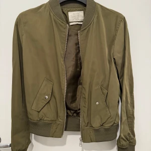 Olivgrön bomberjacka från Bershka - Snygg olivgrön bomberjacka från Bershka med dragkedja och ribbade muddar. Jackan har två fickor med knappar framtill och en klassisk bomberkrage. Perfekt för en stilren look.