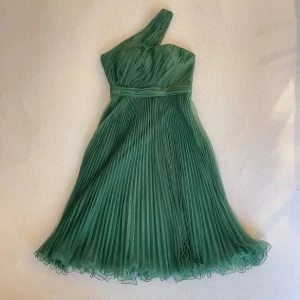 Grön plisserad one shoulder klänning - Säljer en elegant grön klänning med plisserad kjol och enaxlad design. Klänningen har ett vackert fall och markerad midja, perfekt för speciella tillfällen. Tunt, lätt material som ger en luftig känsla.